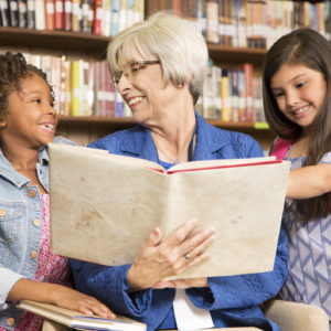 Senior adult teacA senior female volunteer reads a book to elementary students in school classroom or library setting.